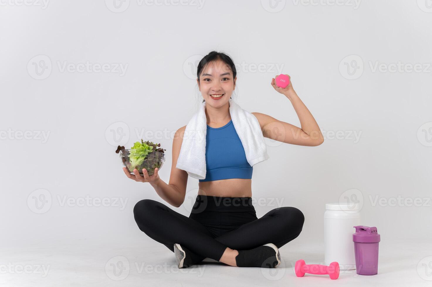 A happy, healthy Asian woman sits on a white studio background, holding a dumbbell and a salad bowl. photo