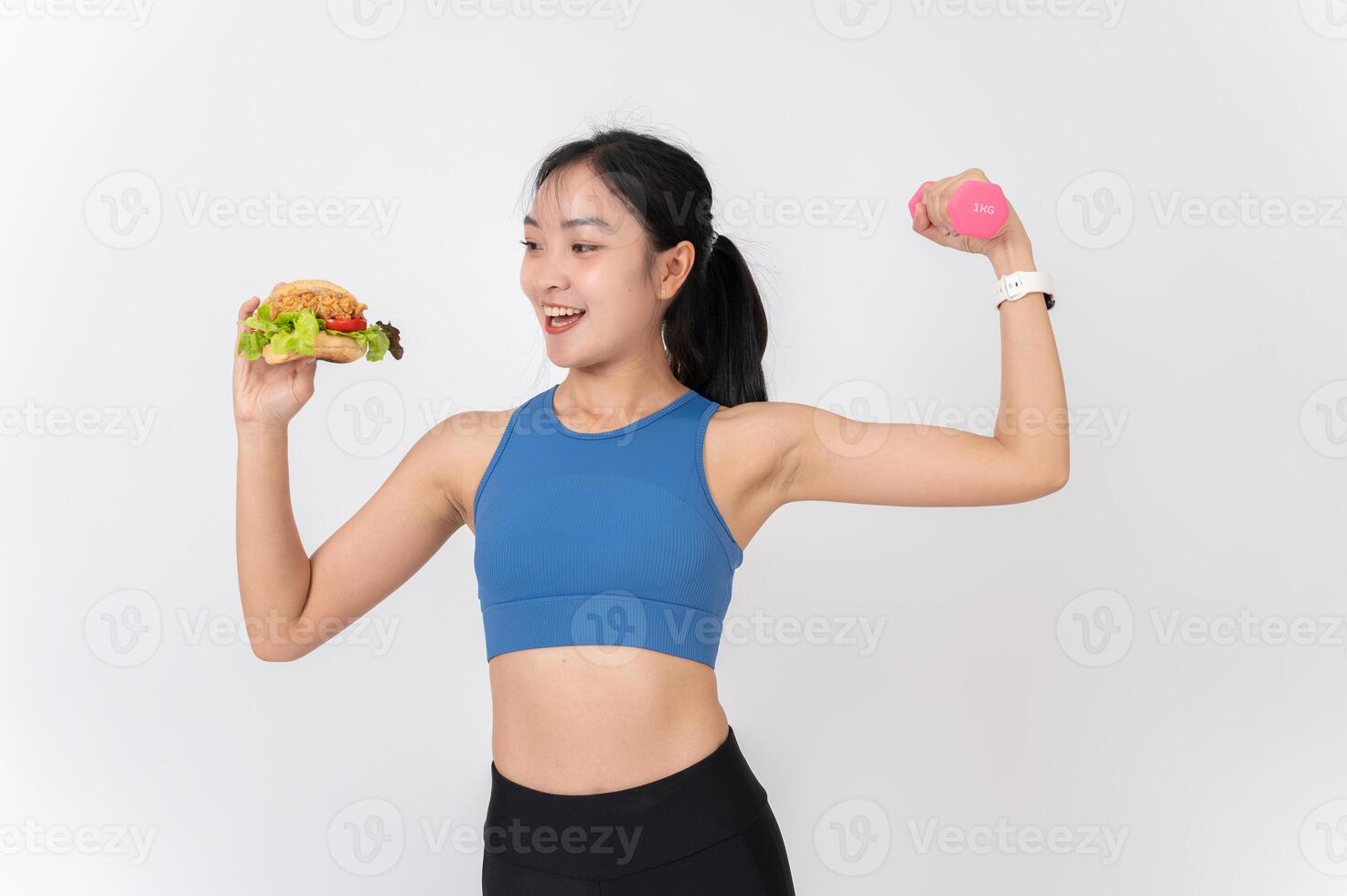 A confident Asian woman in sportswear holding a burger in one hand and a dumbbell in the other. photo