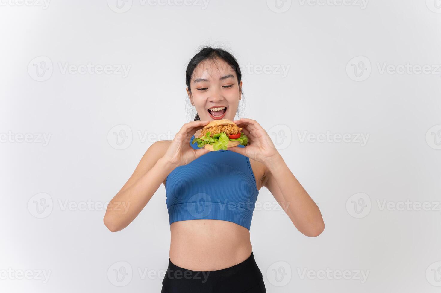 A cheerful Asian woman in sportswear holding a burger, standing against a white studio background. photo