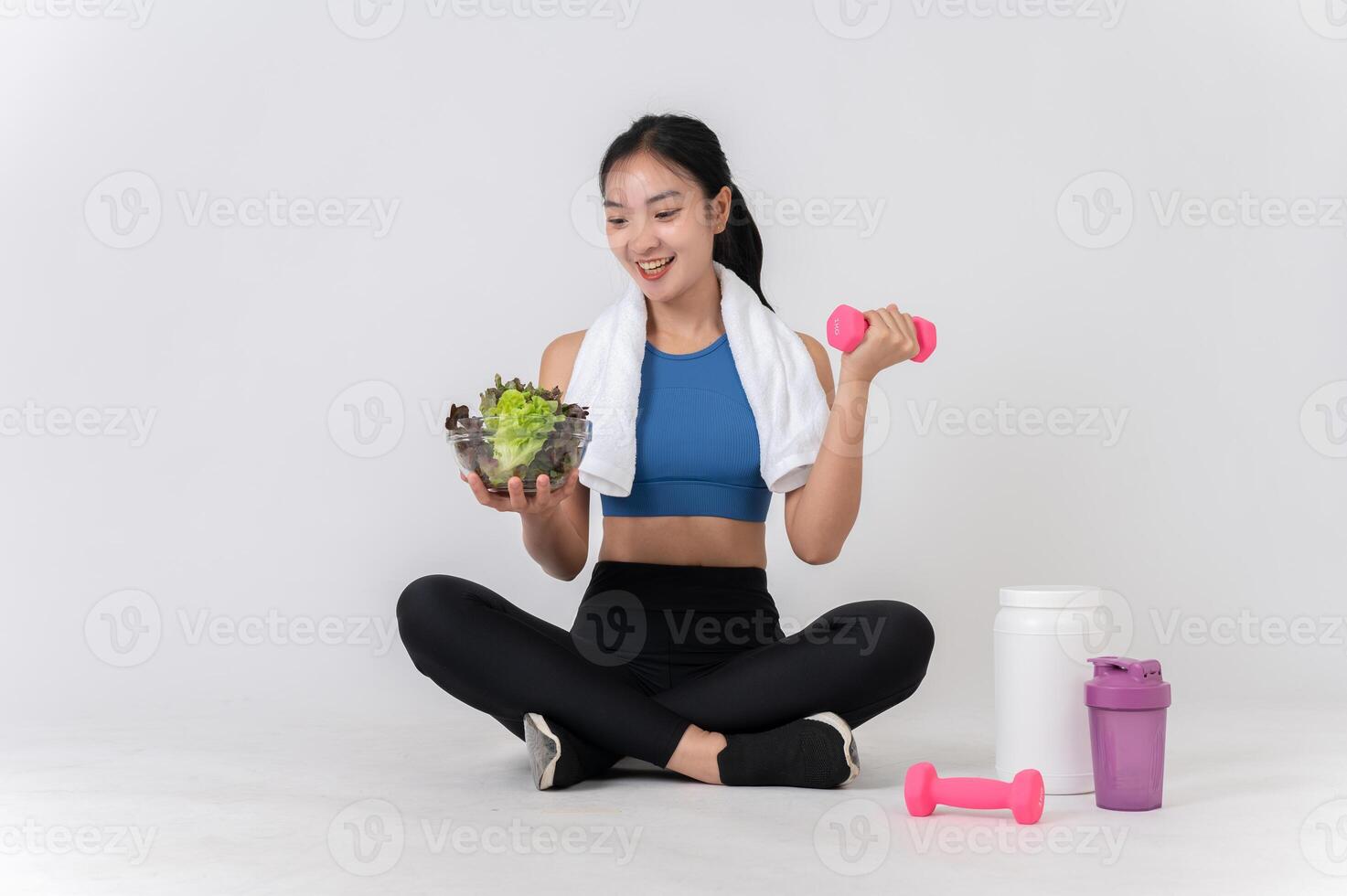 A happy, healthy Asian woman sits on a white studio background, holding a dumbbell and a salad bowl. photo