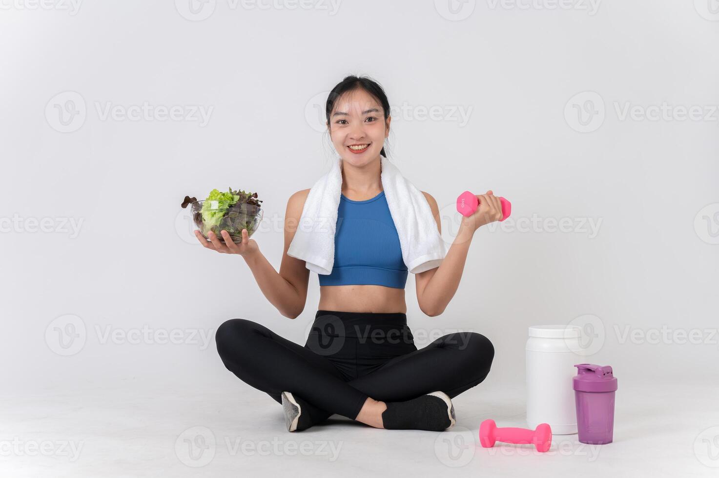 A happy, healthy Asian woman sits on a white studio background, holding a dumbbell and a salad bowl. photo
