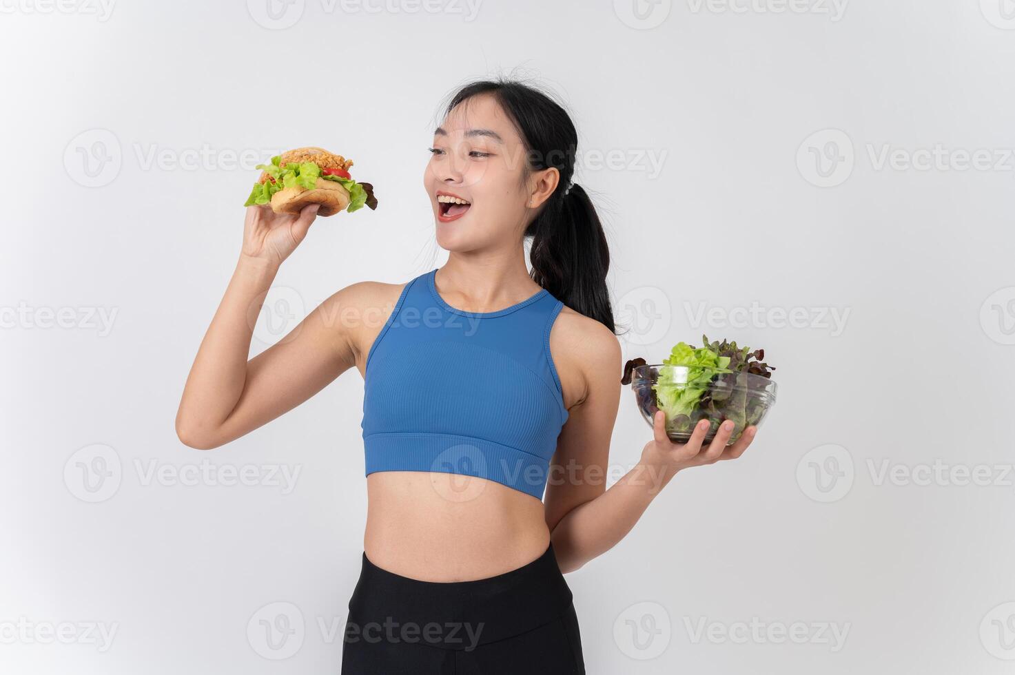 A cheerful Asian woman in sportswear holding a burger in one hand and a salad bowl in the other. photo