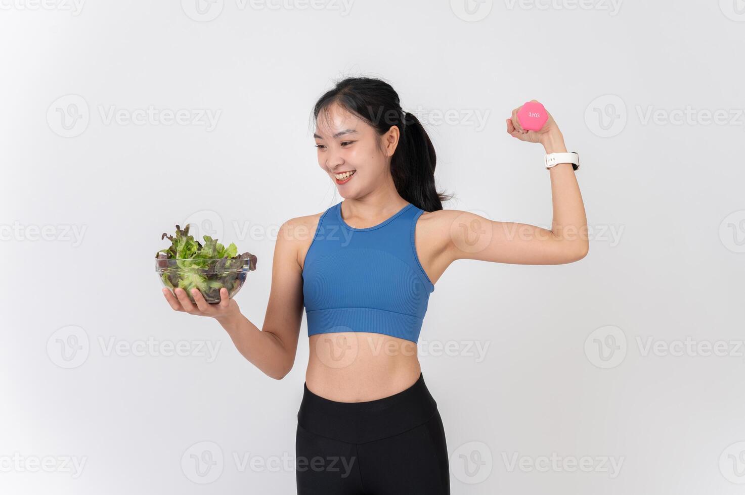 A healthy, confident Asian woman in sportswear holding a salad bowl and a dumbbell. photo