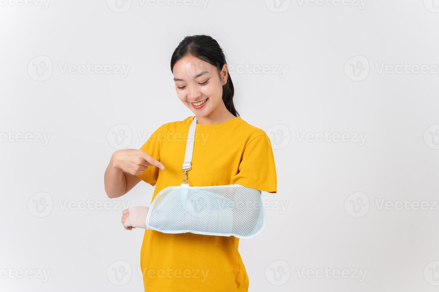 Happy Asian woman pointing at her broken arm, wrapped in a plaster bandage and supported by a sling. photo