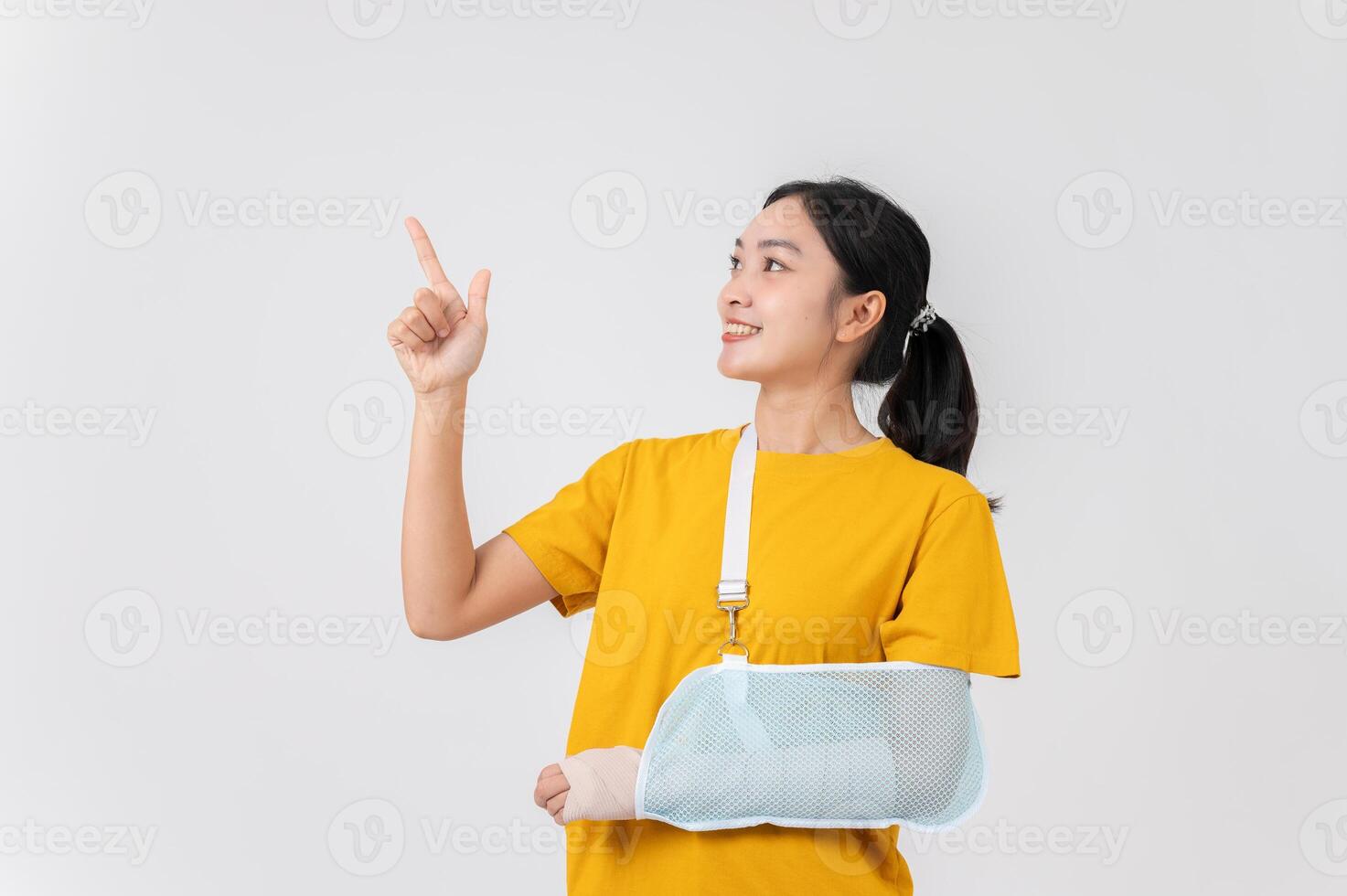 A smiling Asian woman with her broken arm, pointing upward at an empty space, studio background. photo