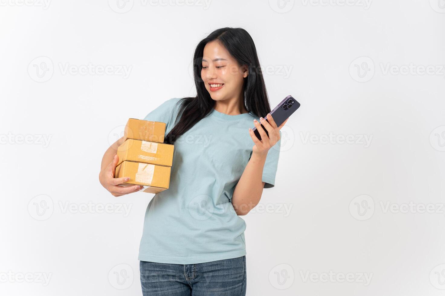 A smiling woman standing against a white studio background, holding cardboard boxes and a smartphone photo
