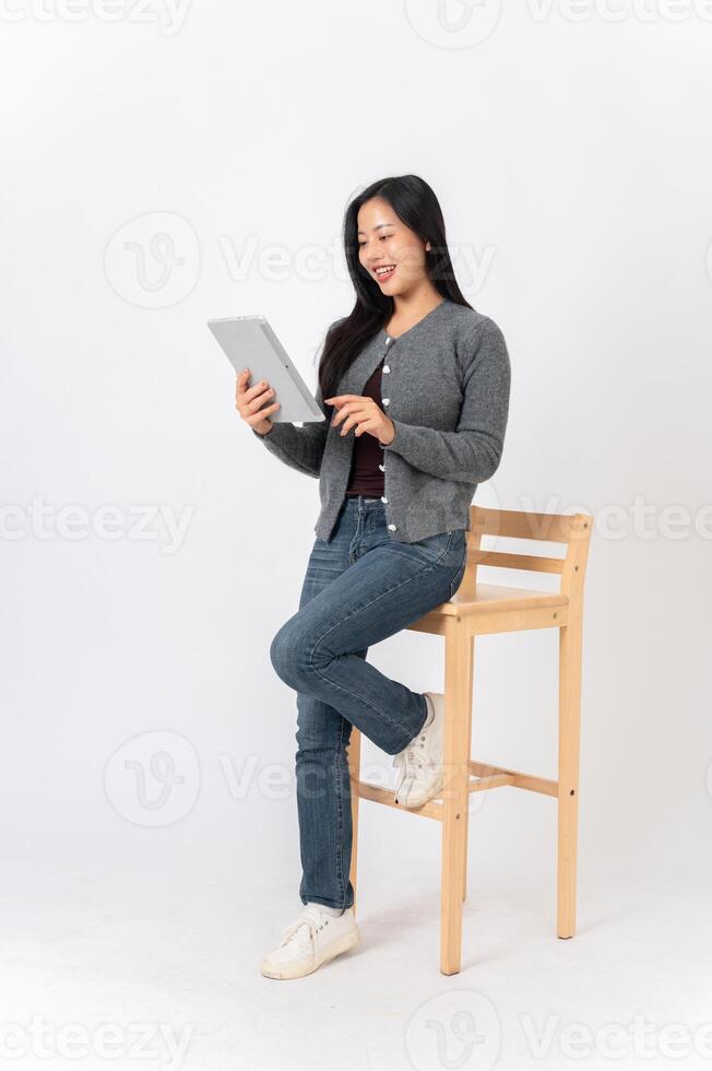 A woman sits on a wooden chair on a white studio background with a digital tablet in her hand. photo