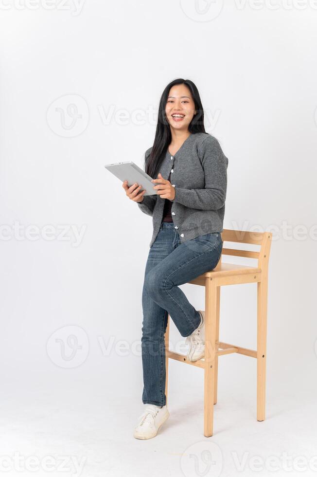 A woman sits on a wooden chair on a white studio background with a digital tablet in her hand. photo