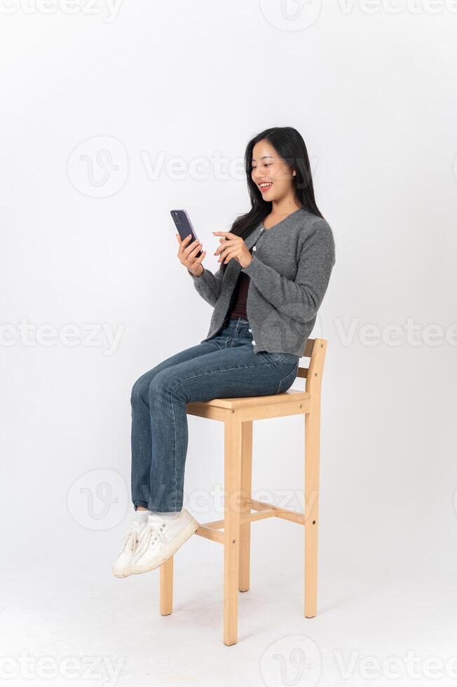 An attractive Asian woman sits on a chair on a white studio background, enjoys using her smartphone. photo