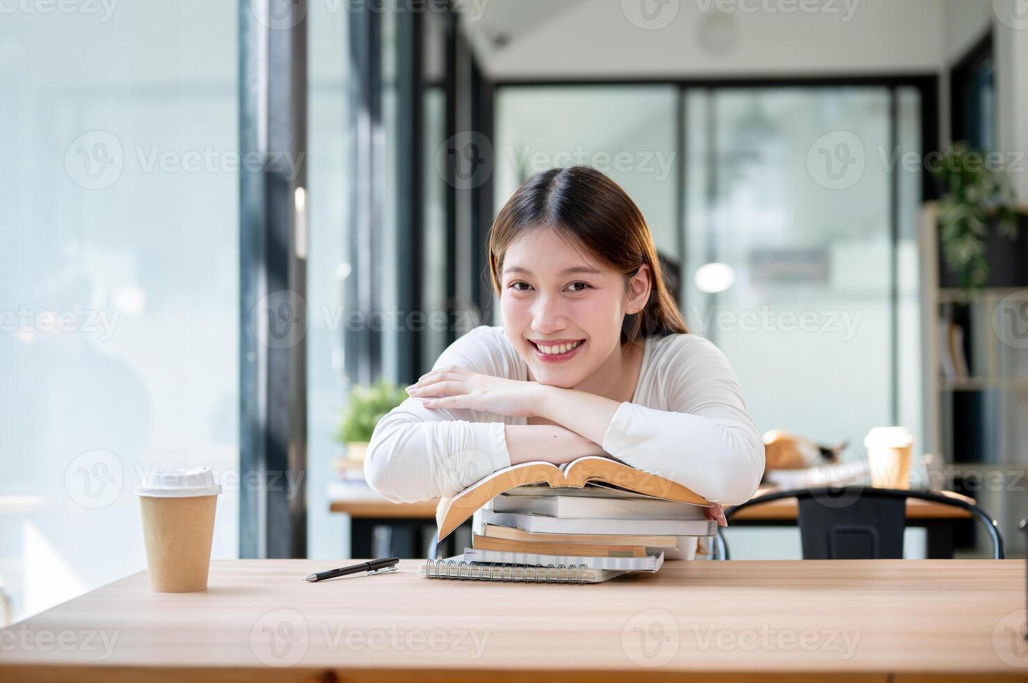 A beautiful Asian woman is resting her hands and face on a stack of books on the table in a library. photo