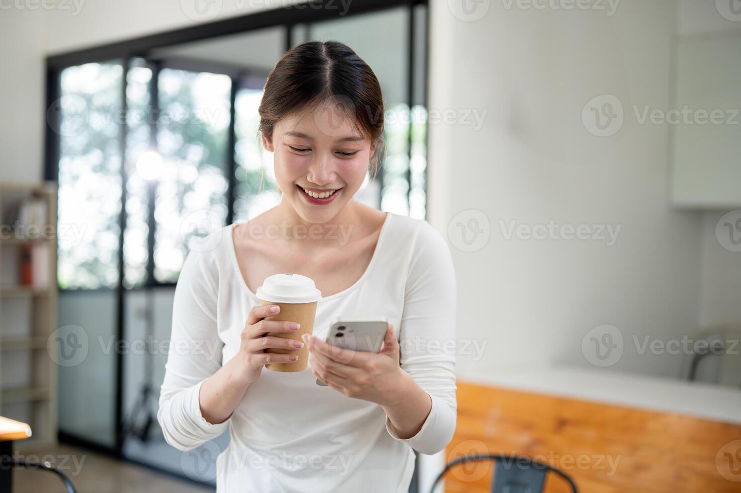 Smiling Asian woman holding a hot drink cup and reading texts on her phone while standing in a cafe. photo