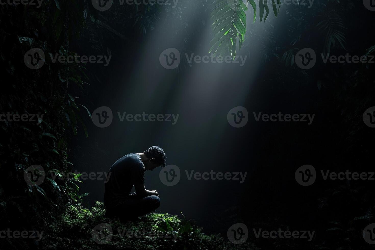 Man kneeling in a dark forest with beams of light breaking through trees photo