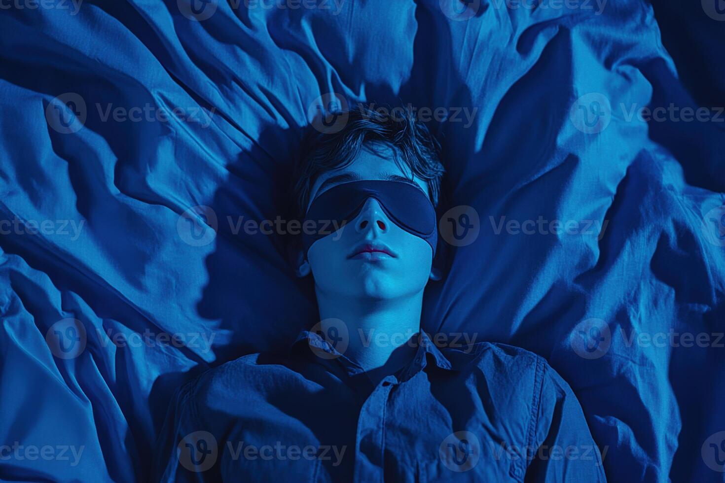 Teenager resting on blue sheets while wearing an eye mask in dim light photo