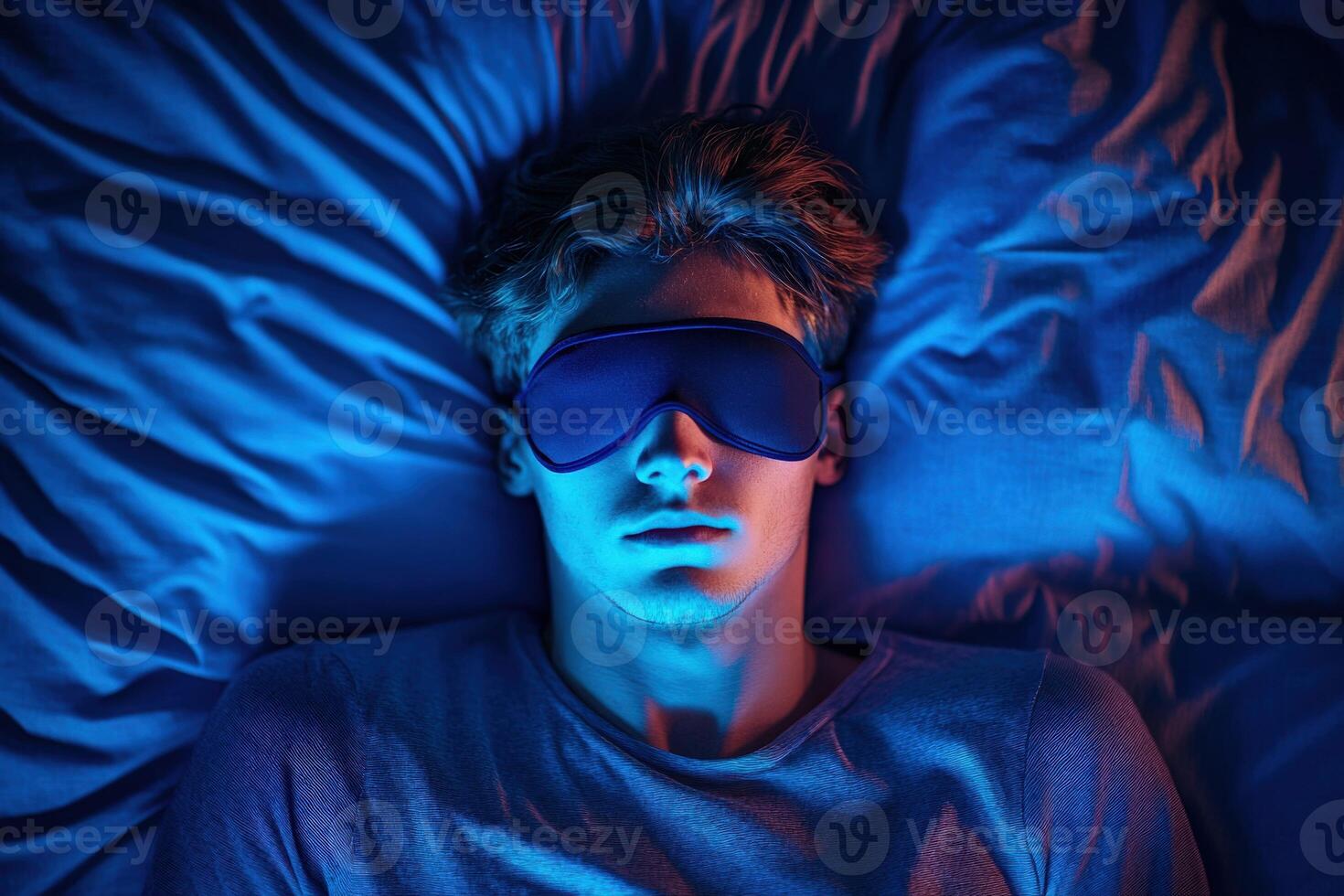 Young man relaxing in bed with sleep mask under blue light at night photo