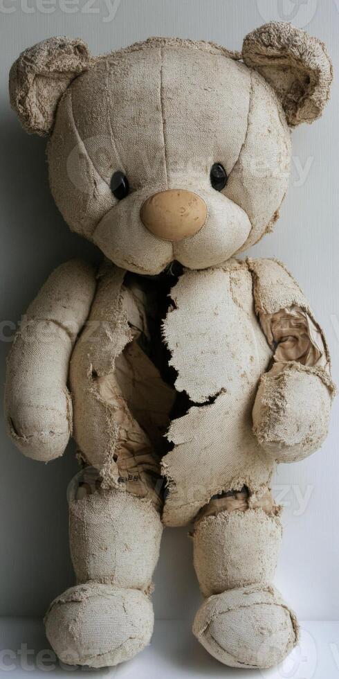 Worn teddy bear with tattered fur and exposed stuffing sits against a wall photo