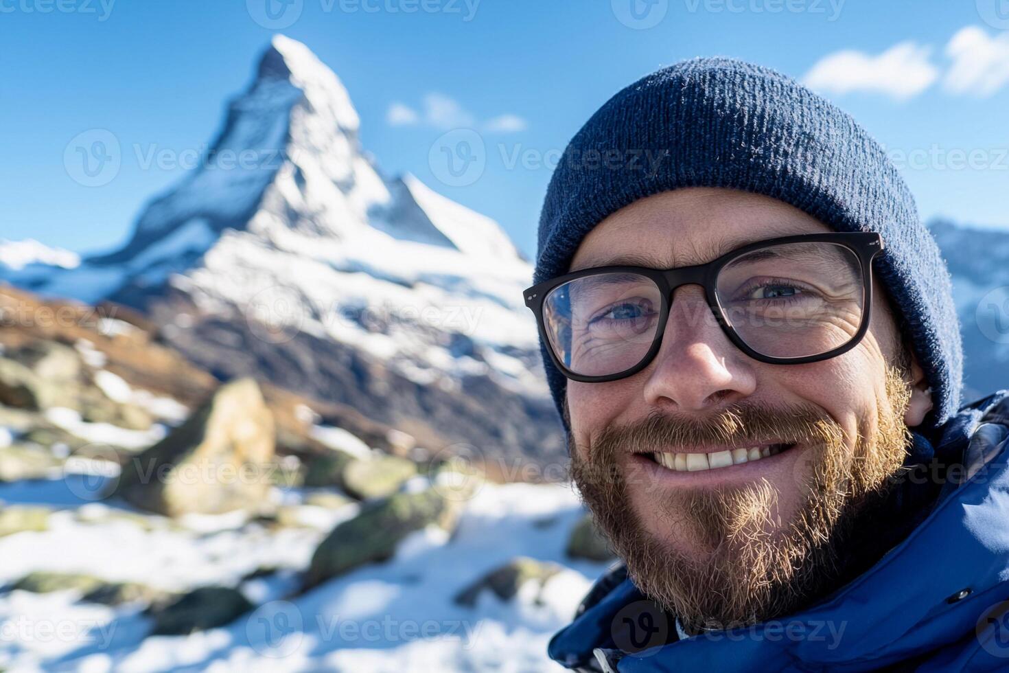 A man with glasses and a blue hat is smiling in front of a mountain photo