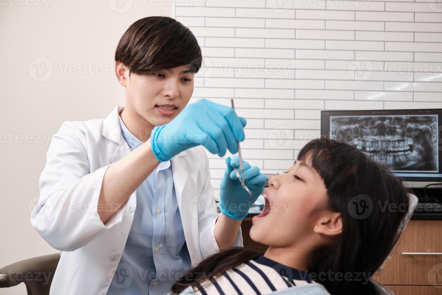A young Asian male pediatric dentist checks and examines a little Thai girl's teeth in dental clinic, well-being mouth oral hygiene, and professional orthodontic healthcare work in a kid hospital. photo