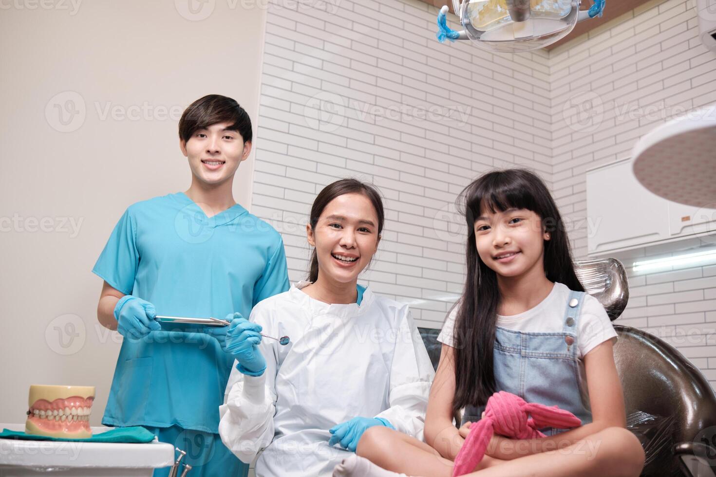 Portrait of Asian female pediatric dentist, assistant, and a girl patient smiling and looking at camera in dental clinic, mouth hygiene, and professional orthodontic healthcare work in a kid hospital. photo