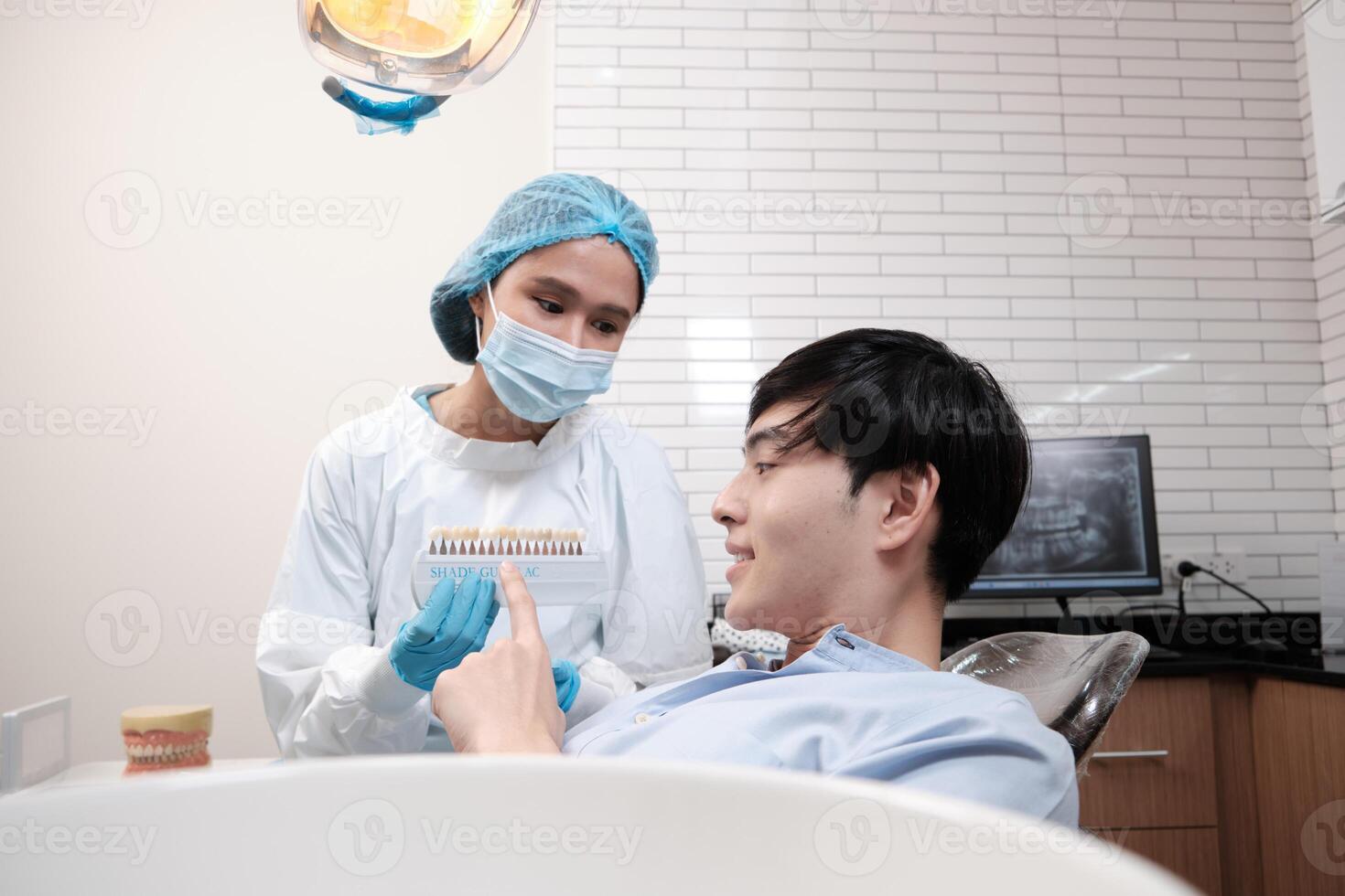 Asian female dentist explains color shades of tooth implant whitening to young male patient, look at mirror in dental clinic, and do professional mouth hygiene healthcare work in a doctor's hospital. photo