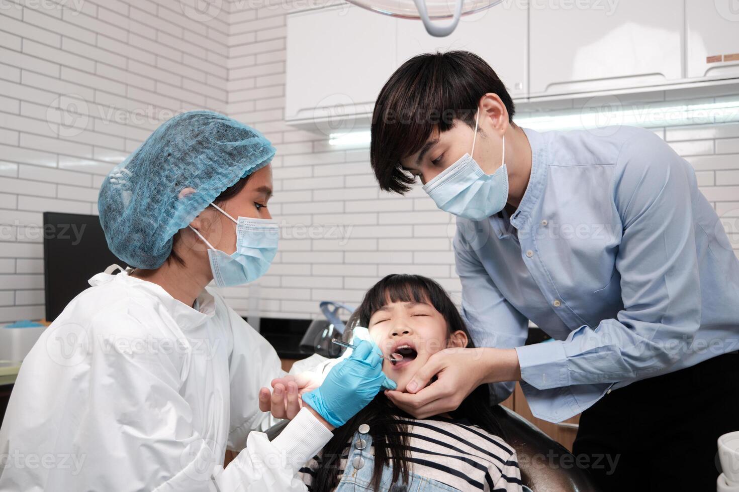 Asian female pediatric dentist examines a girl's teeth in dental clinic and takes care and encourages of father, mouth oral hygiene, and professional orthodontic healthcare work in a kid hospital. photo