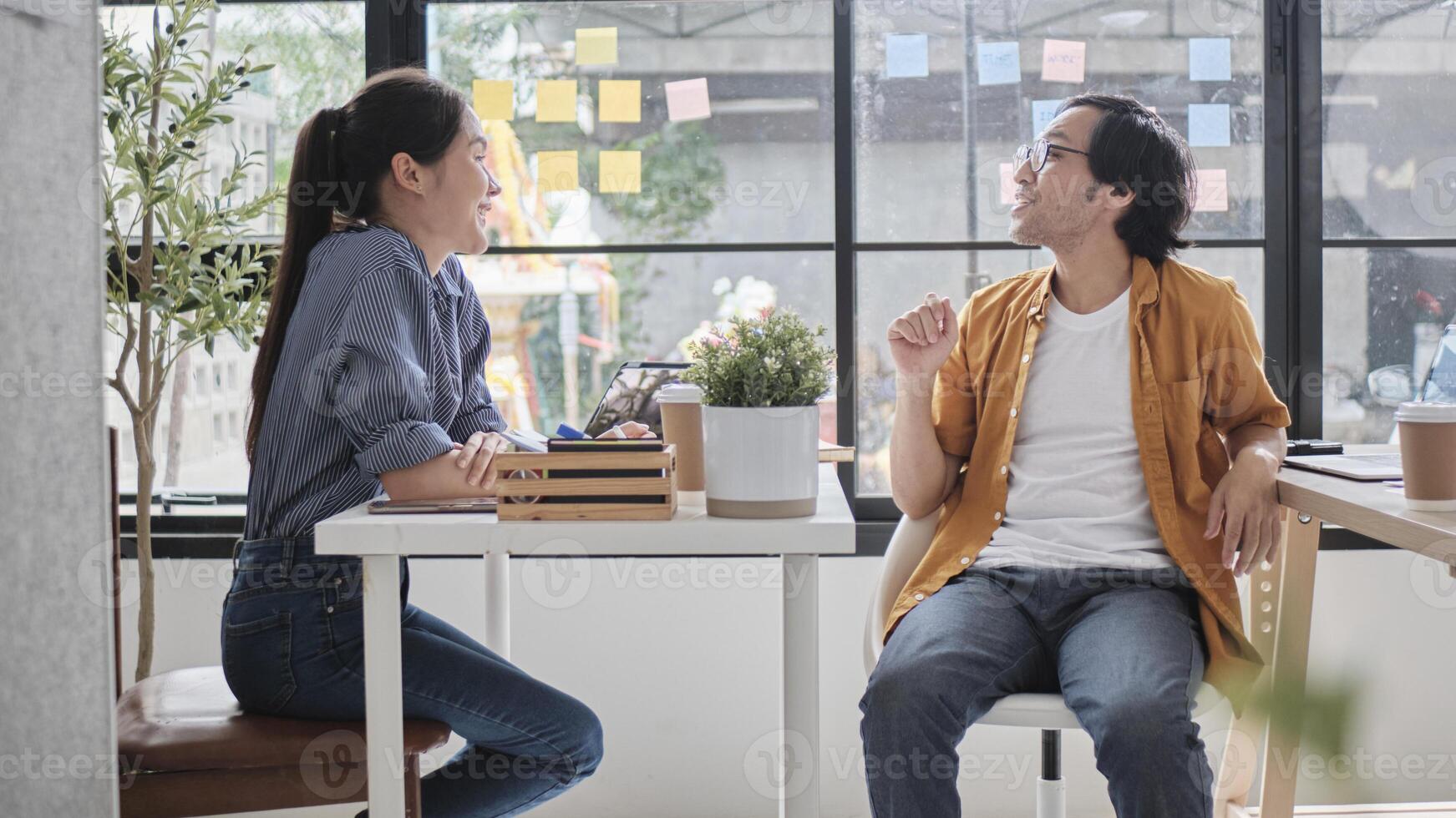 Two Asian workers sitting in chairs at working desks, collaborating and talking at daylight windows in casual small business office, online business job coworkers, positive relationship colleagues. photo