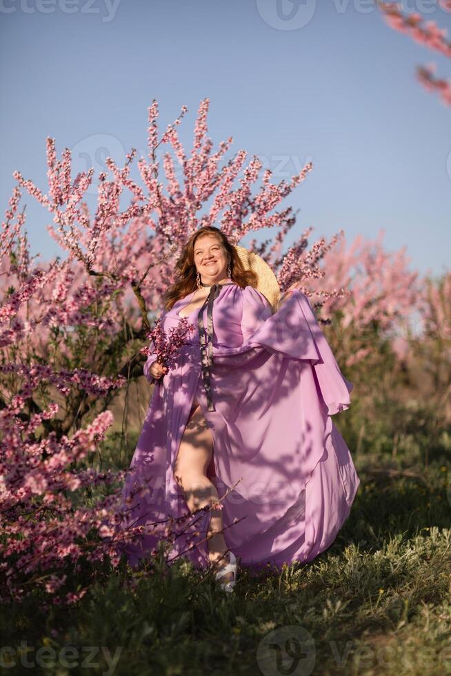Woman blooming peach orchard. Against the backdrop of a picturesque peach orchard, a woman in a long pink dress and hat enjoys a peaceful walk in the park, surrounded by the beauty of nature. photo