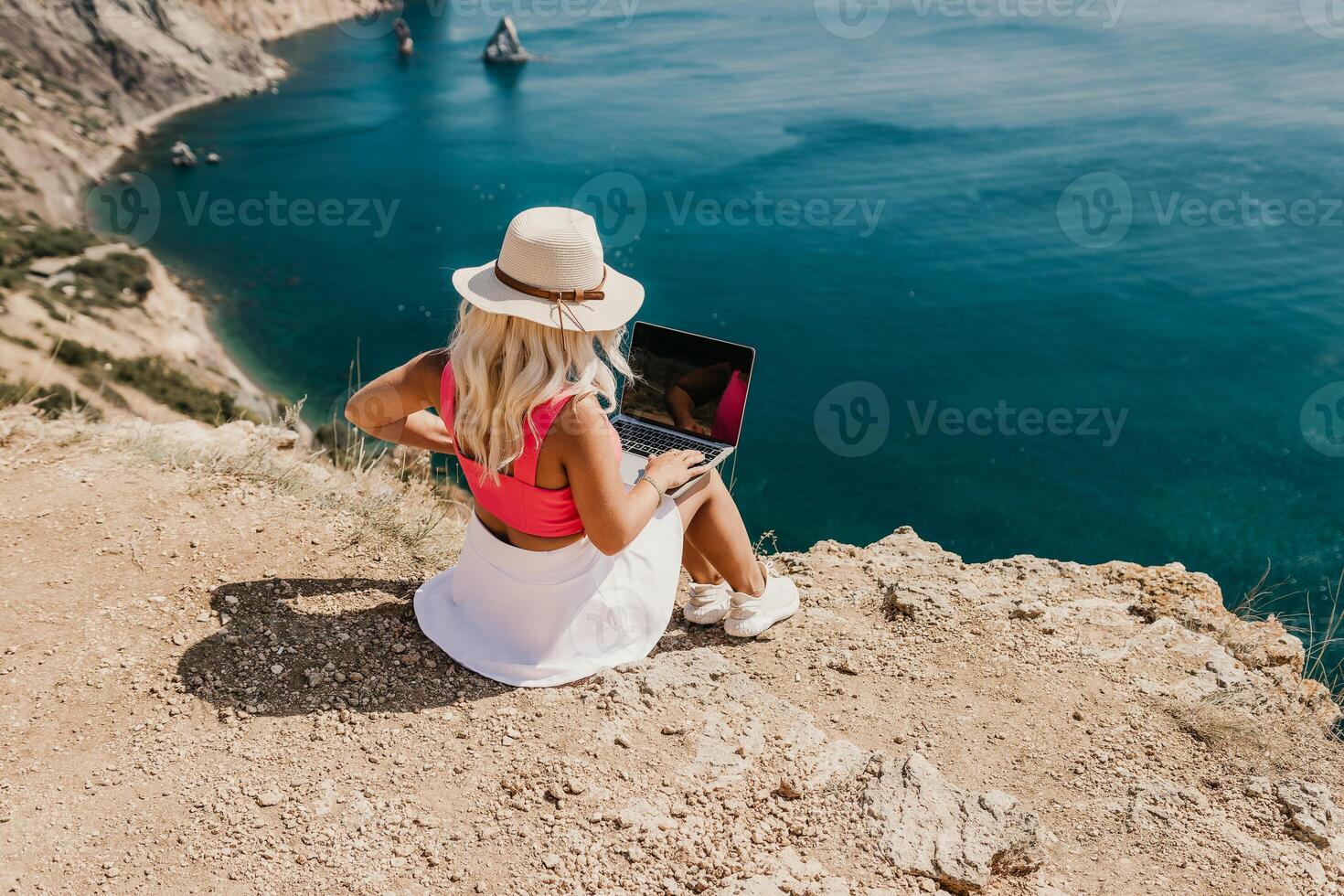 A woman is sitting on a rocky beach with a laptop open in front of her. She is wearing a pink top and a straw hat. Concept of relaxation and leisure. photo