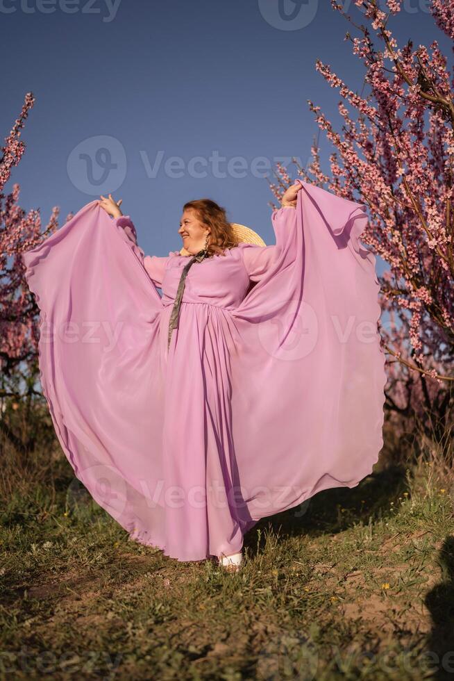 Woman blooming peach orchard. Against the backdrop of a picturesque peach orchard, a woman in a long pink dress and hat enjoys a peaceful walk in the park, surrounded by the beauty of nature. photo