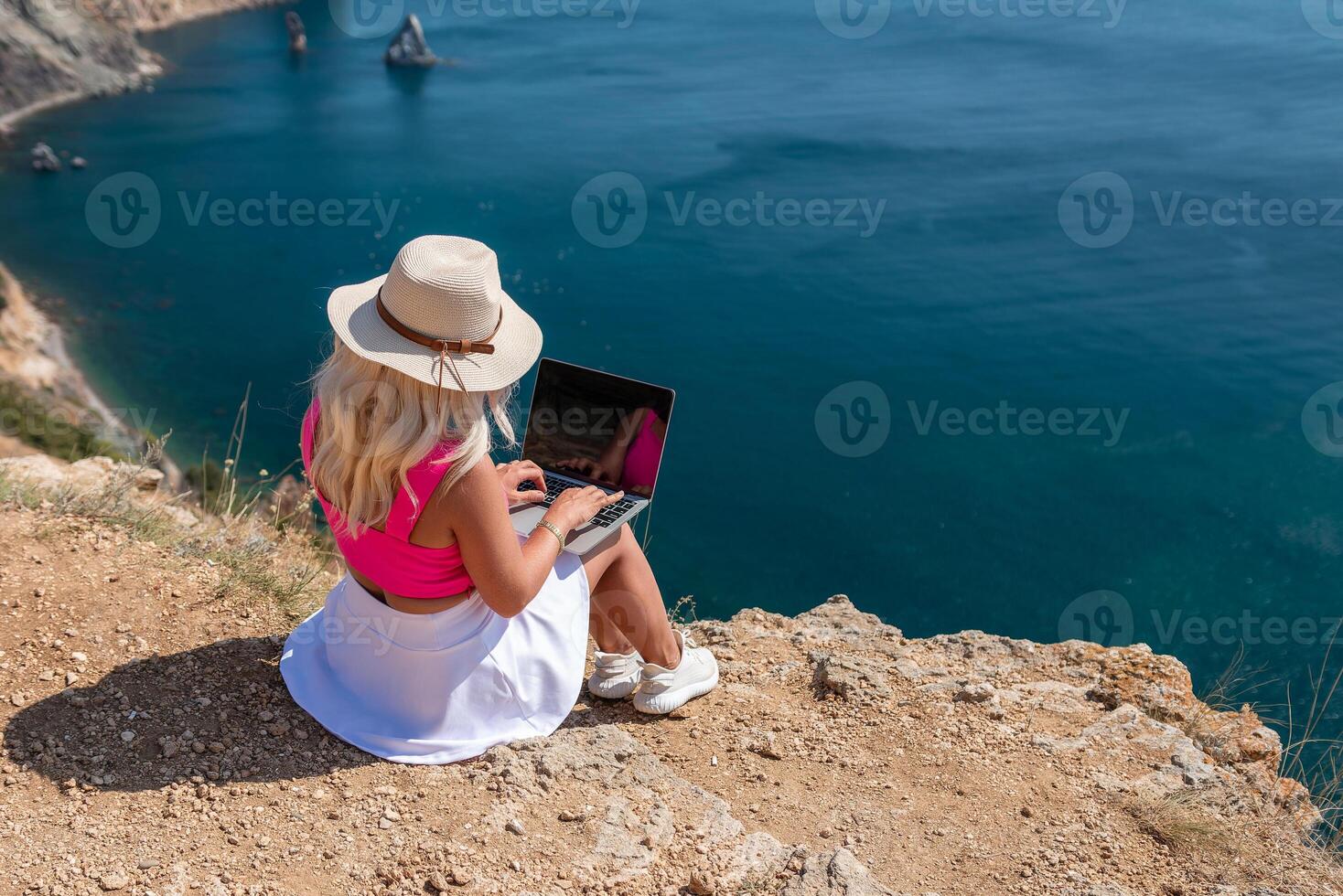 A woman is sitting on a rocky beach with a laptop open in front of her. She is wearing a pink top and a straw hat. Concept of relaxation and leisure. photo