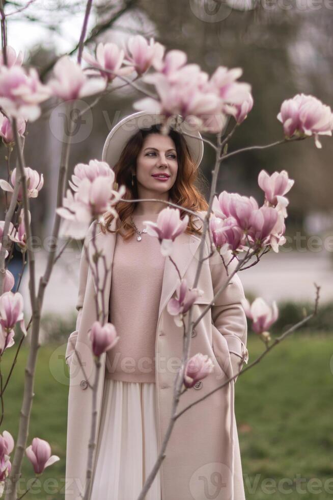 Woman magnolia flowers, surrounded by blossoming trees., hair down, white hat, wearing a light coat. Captured during spring, showcasing natural beauty and seasonal change. photo