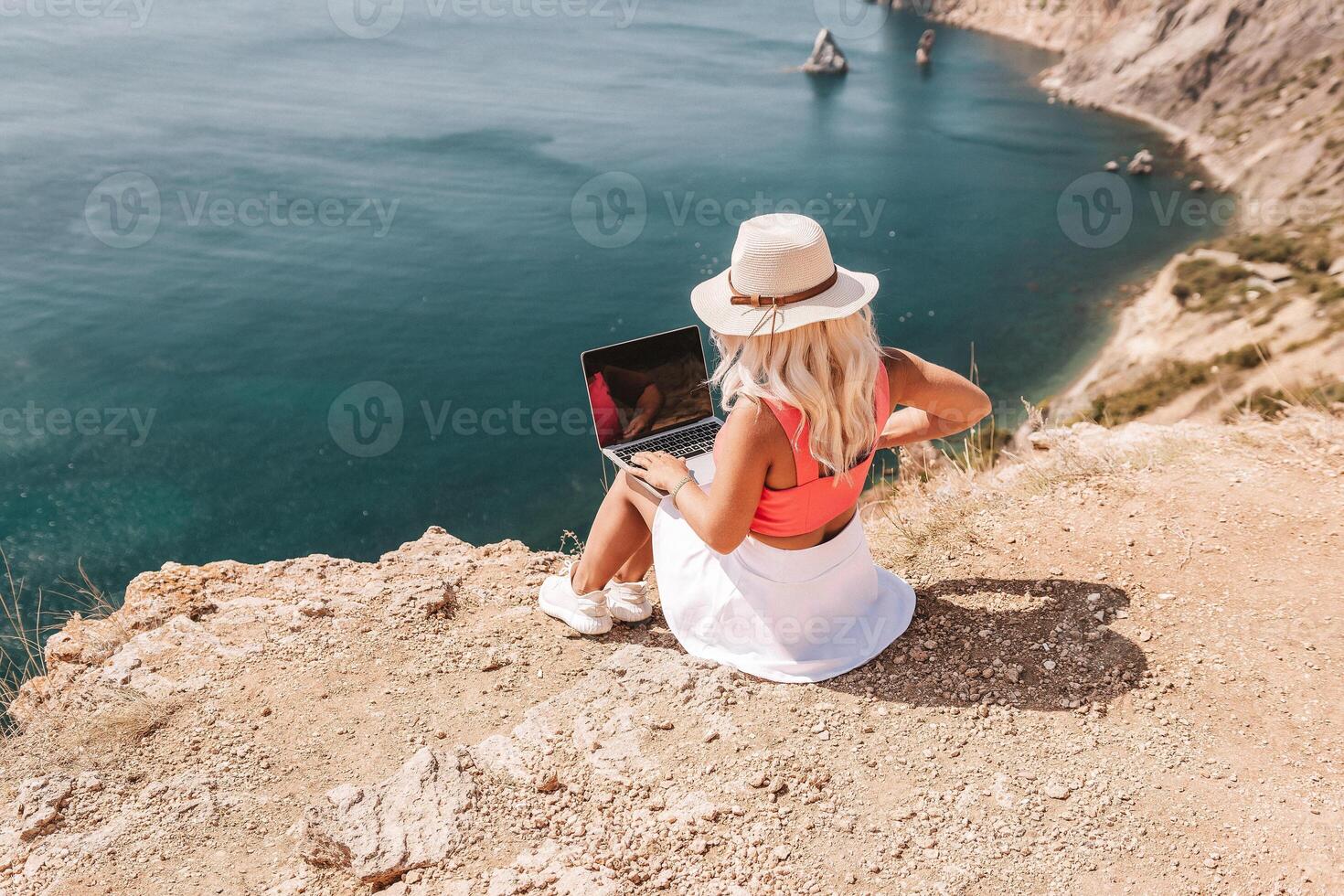 A woman is sitting on a rocky beach with a laptop open in front of her. She is wearing a pink top and a straw hat. Concept of relaxation and leisure. photo