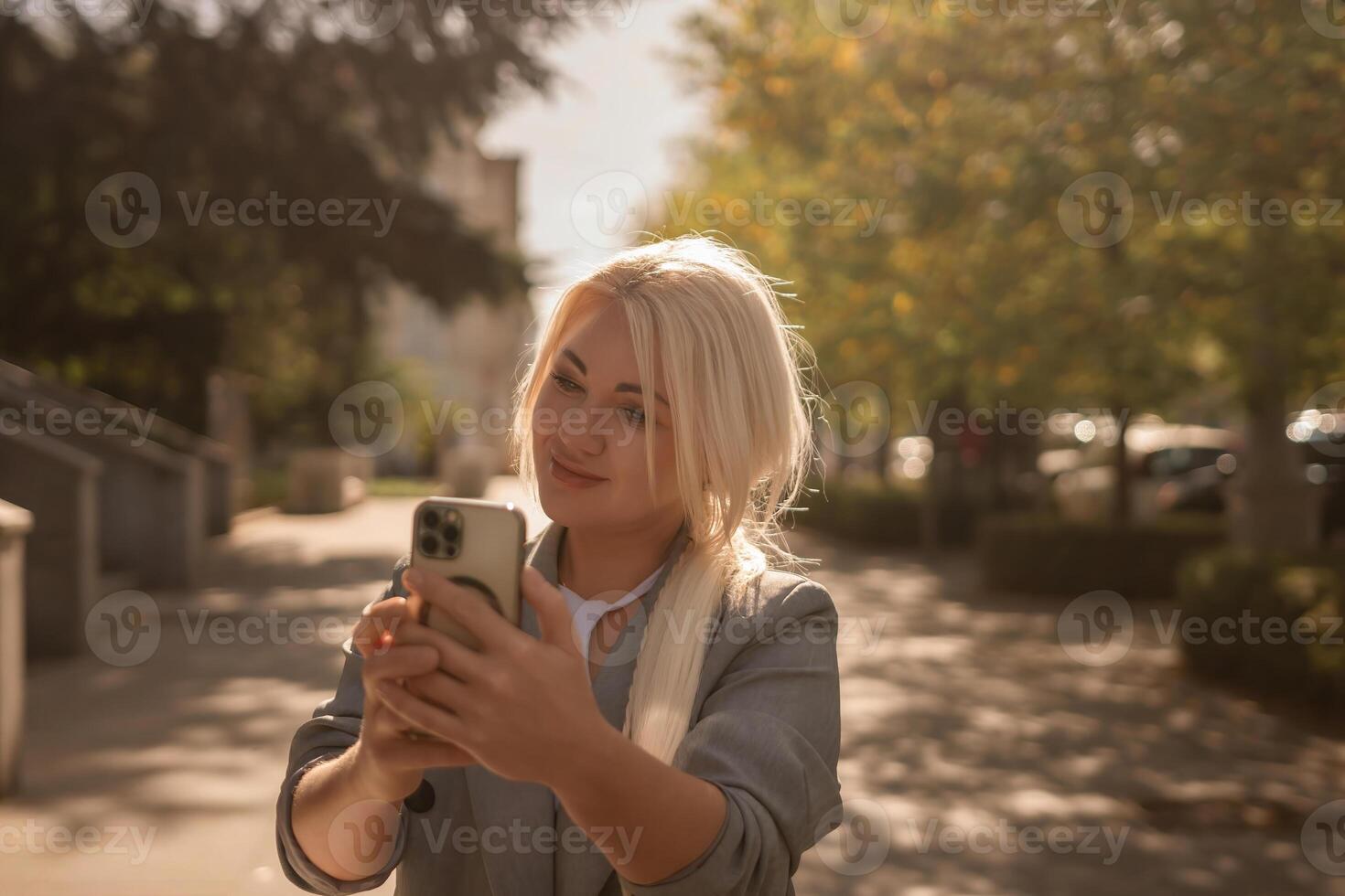 A woman is taking a picture of herself with her cell phone. She is wearing a gray jacket and scarf. The scene is set in a city with trees and cars in the background. photo