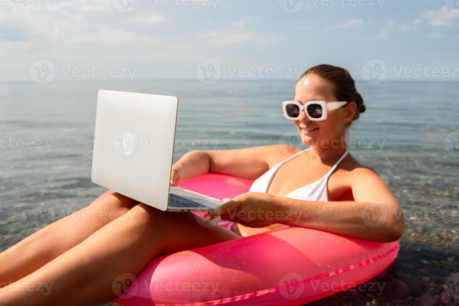 A woman is sitting on a pink inflatable raft with a laptop on her lap. She is wearing sunglasses and a white bikini top. Concept of relaxation and leisure. photo