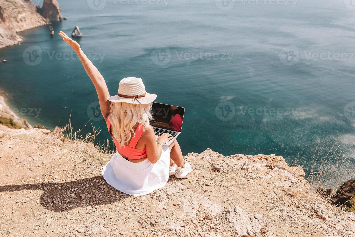 A woman is sitting on a rocky beach with a laptop in front of her. She is wearing a pink top and a straw hat. She is waving her hand in the air, possibly to celebrate or to show excitement. photo