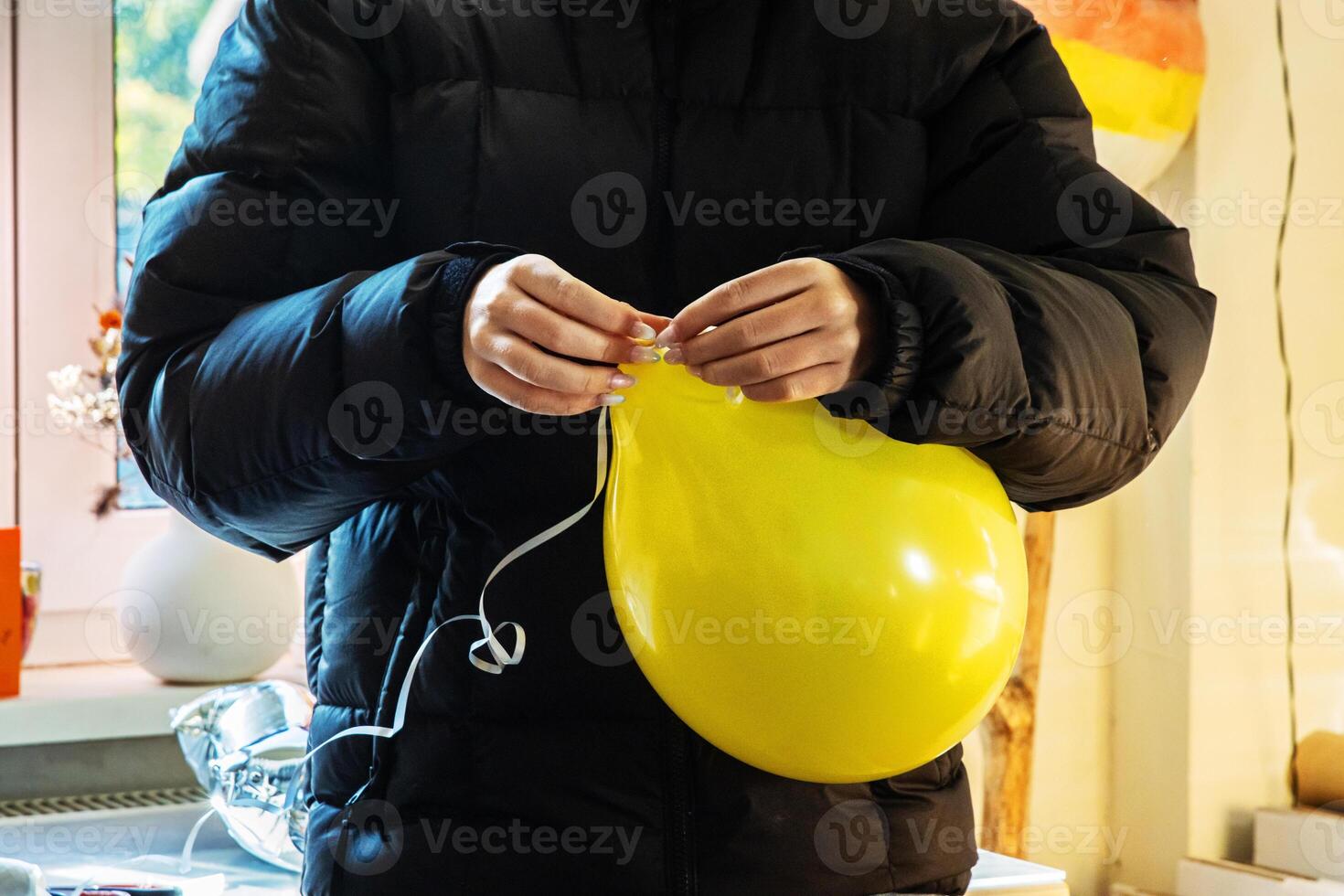 Teenager girl in a black jacket preparing decorations for a birthday celebration at home by inflating balloons and tying a string to a yellow balloon photo