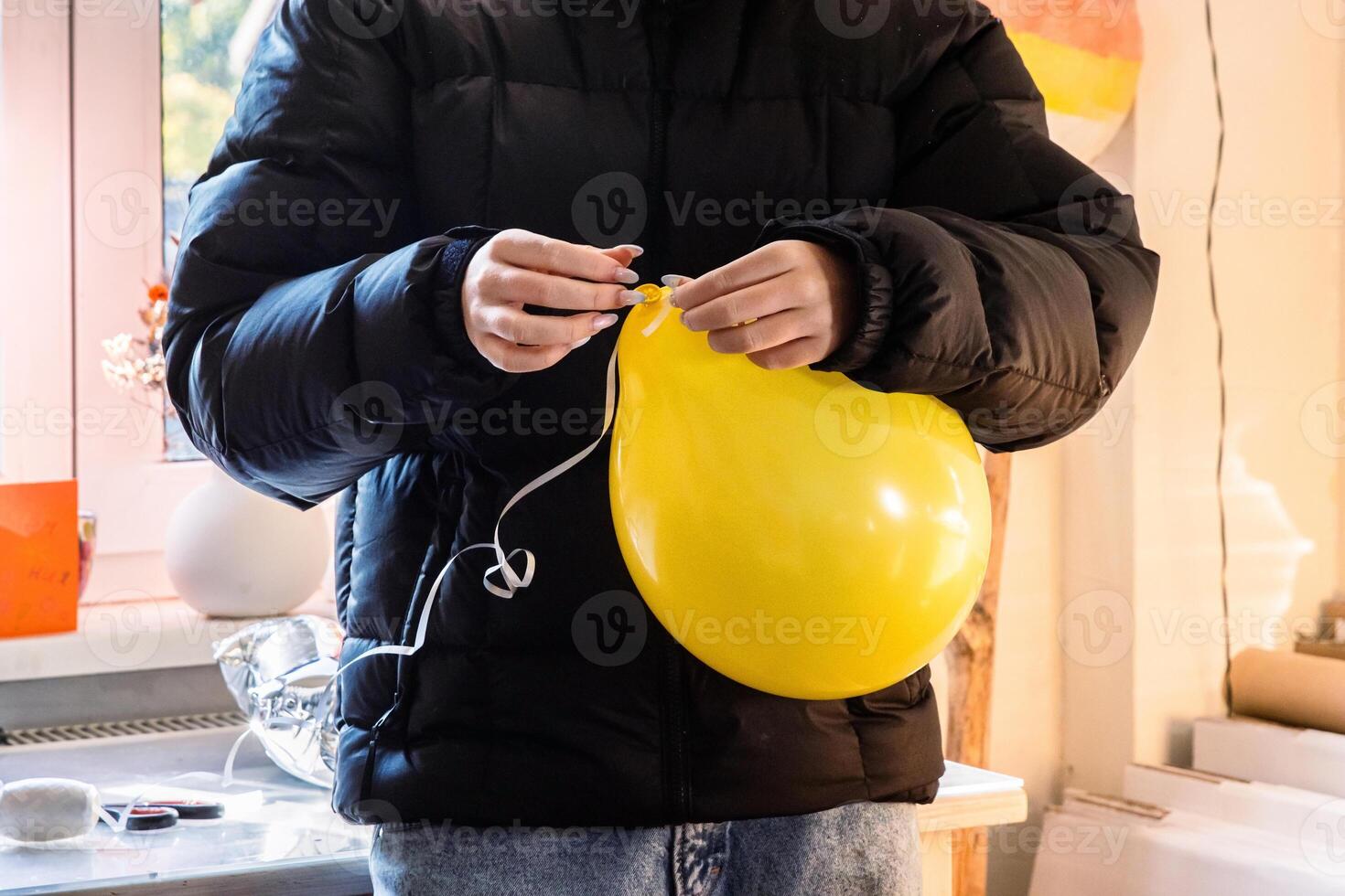 Teenager girl in a black jacket preparing decorations for a birthday celebration at home by inflating balloons and tying a string to a yellow balloon. photo
