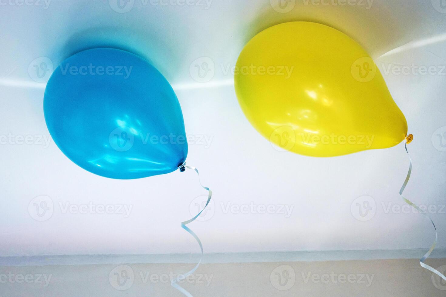 Two bright helium balloons in contrasting yellow and blue colors tied to ribbons, floating under a ceiling, creating a cheerful atmosphere at children's birthday celebration. Copy space photo