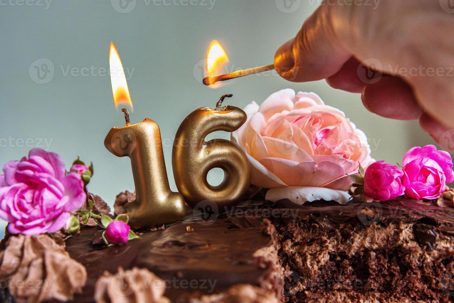 Closeup of a womans hand lighting golden 16 candles on a chocolate birthday cake decorated with pink and peach roses for a sweet sixteen celebration photo