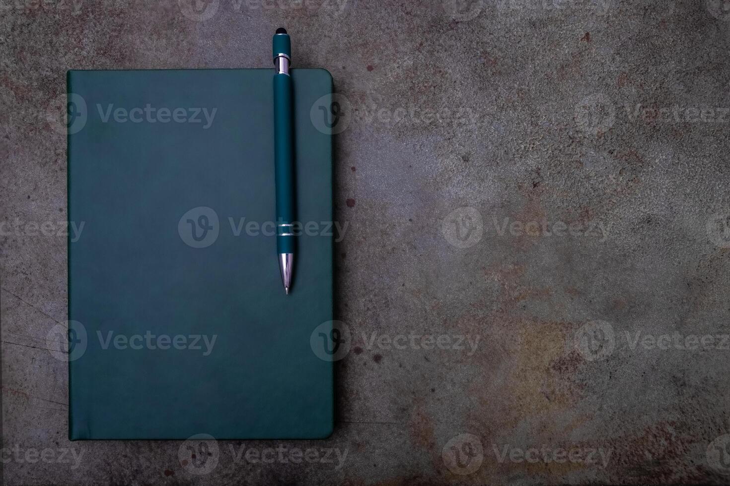 A green bound notebook on a light brown, textured surface, accompanied by a pen photo