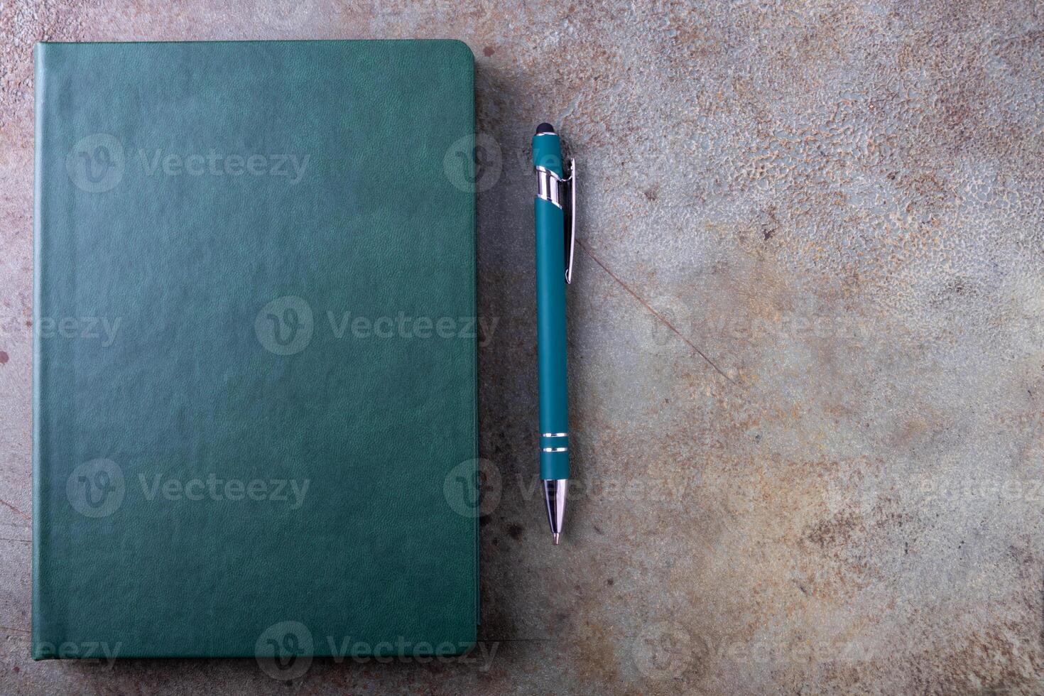 A green bound notebook on a light brown, textured surface, accompanied by a pen photo