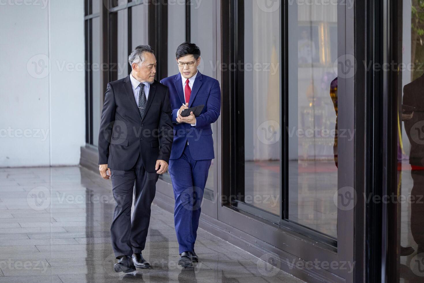 Asian senior businessman giving advice to the new manager and looking at information regarding sale record while walking along modern style office photo