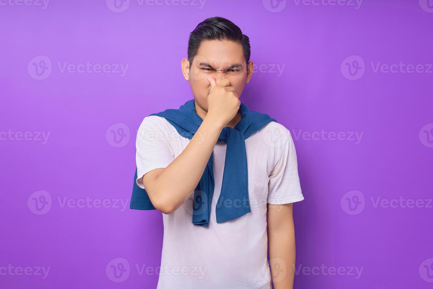 confused young Asian man wearing white t-shirt looking at the camera and covering his nose with a hand isolated over purple background. people lifestyle concept photo