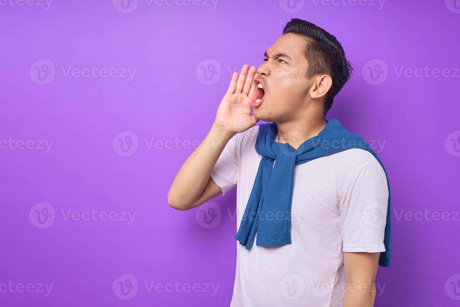 Excited young Asian man wearing white t-shirt screaming with hands gesture near his mouth looking aside isolated over purple background. people lifestyle concept photo