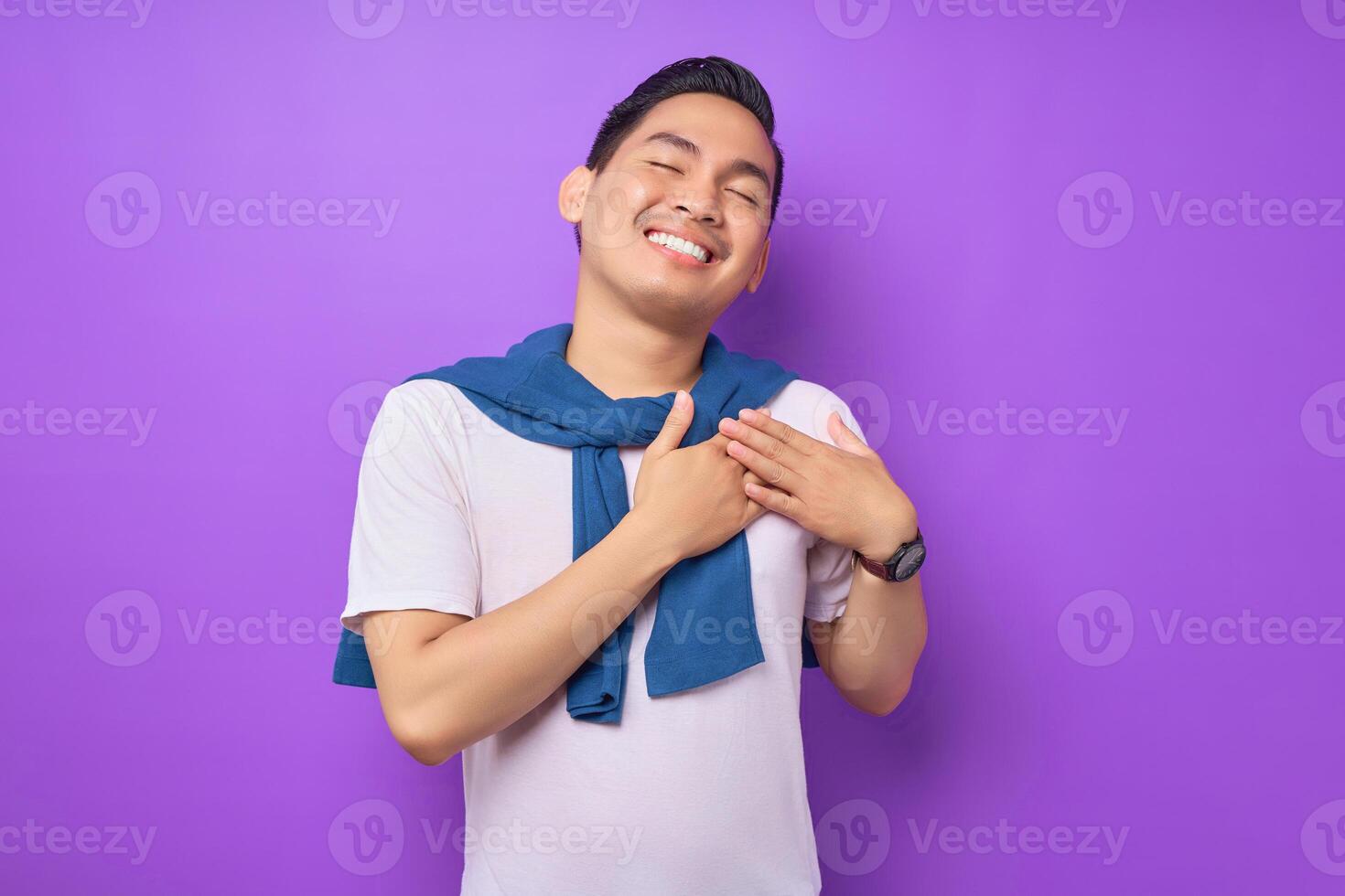Enjoy angry young Asian man wearing white t-shirt keeping his eyes closed, holding palms crossed on his chest isolated over purple background. people lifestyle concept photo