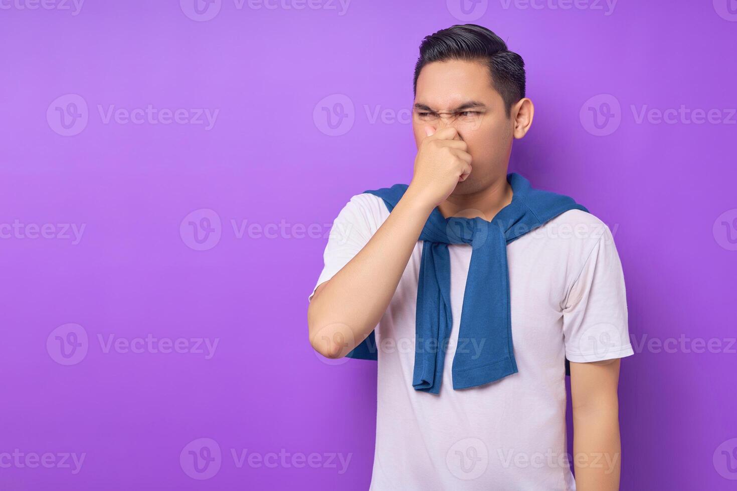 disconcerted young Asian man wearing white t-shirt looking aside covering nose with hand isolated over purple background. people lifestyle concept photo