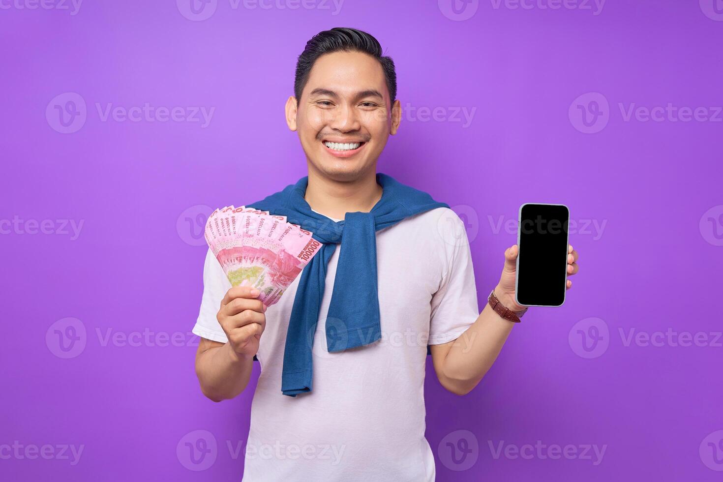 Cheerful young Asian man holding mobile phone with blank screen and money banknotes isolated over purple background. Easy online money transfer concept photo