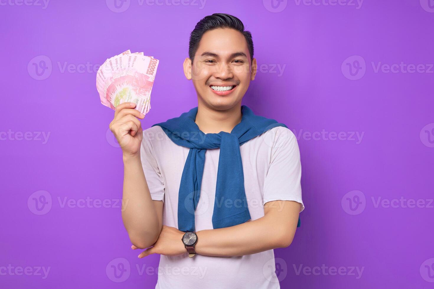 Happy young Asian man wearing white t-shirt casual clothes holding money banknotes and smiling at camera isolated over purple background. Profit and wealth concept photo