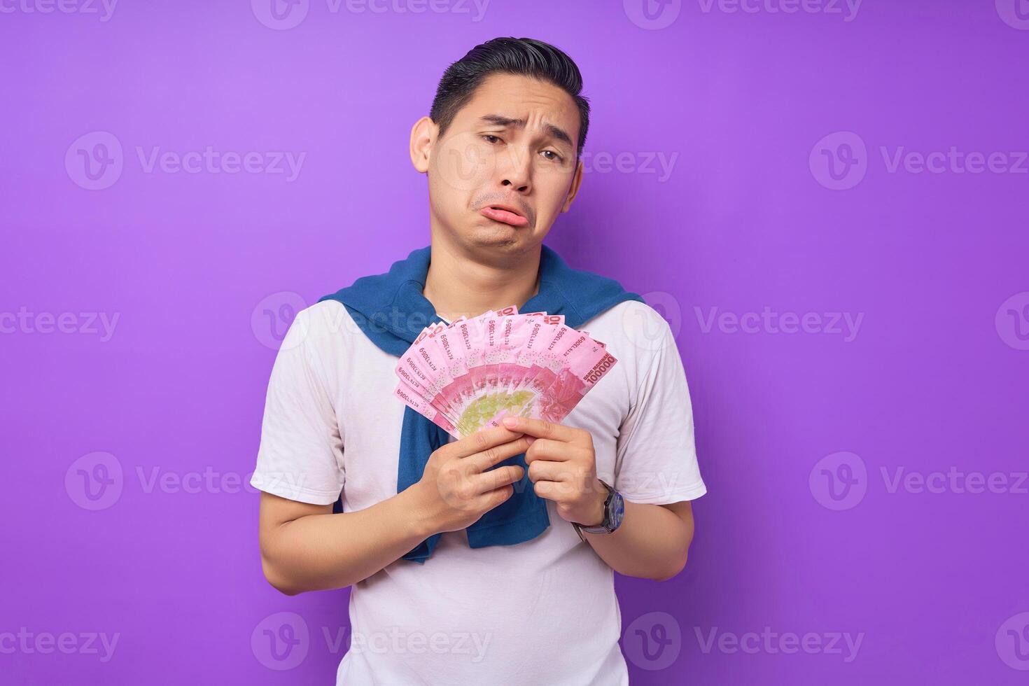 Sad young Asian man wearing white t-shirt holding money banknotes and looking at camera isolated over purple background. Profit and wealth concept photo