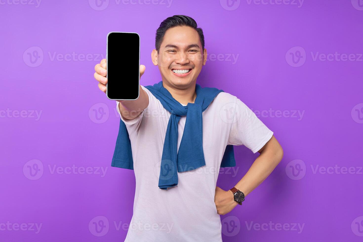 Smiling young Asian man holding smartphone with blank screen isolated on purple background. Advertising new mobile app, mockup concept photo