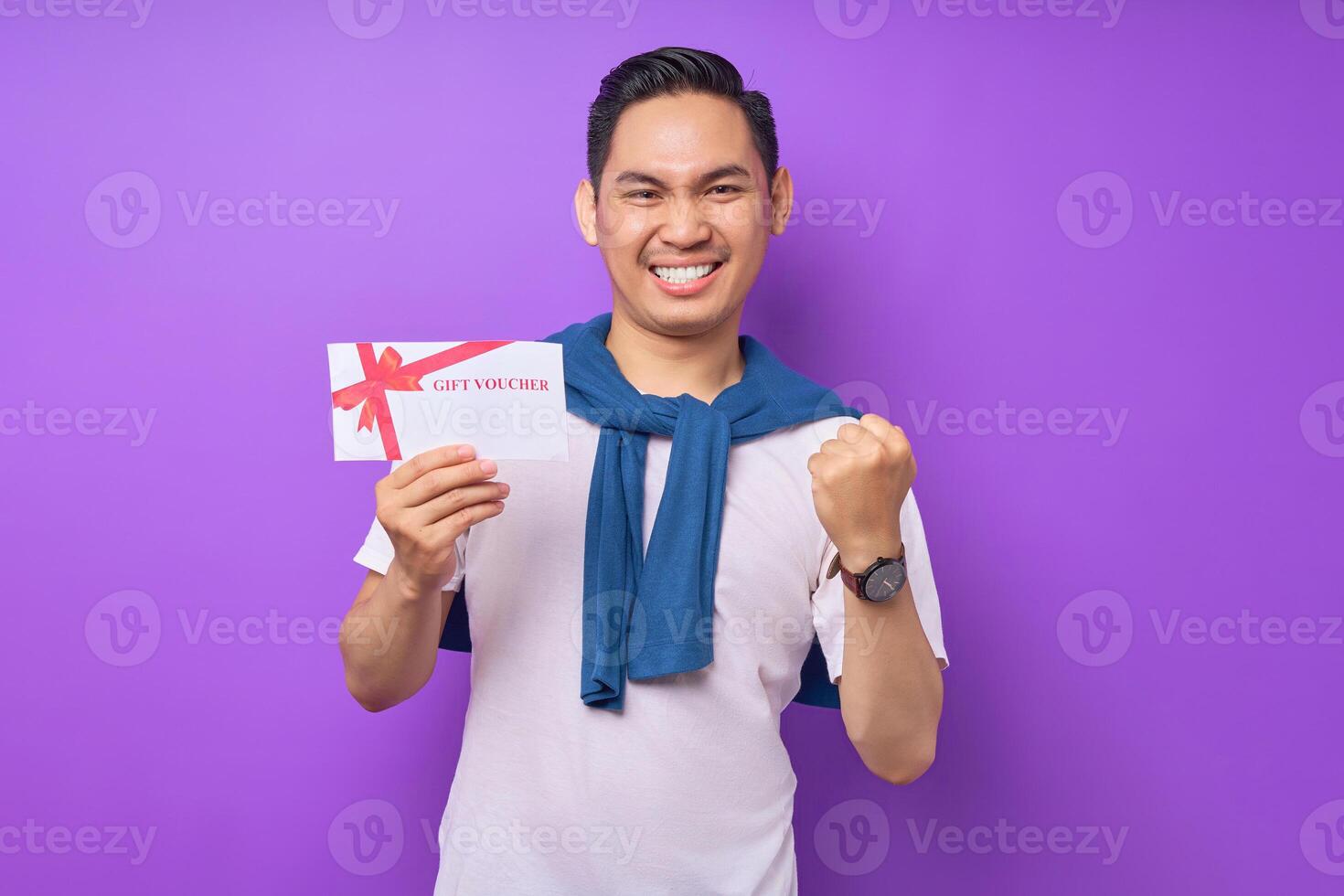 Smiling young Asian man in casual clothes holding a gift voucher certificate and celebrating winner isolated on purple studio background photo
